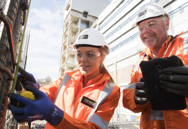 Two Laing O'Rourke Australia employees working on a construction site. The person on the right is holding a tablet, the person on the left is using a measuring tape.